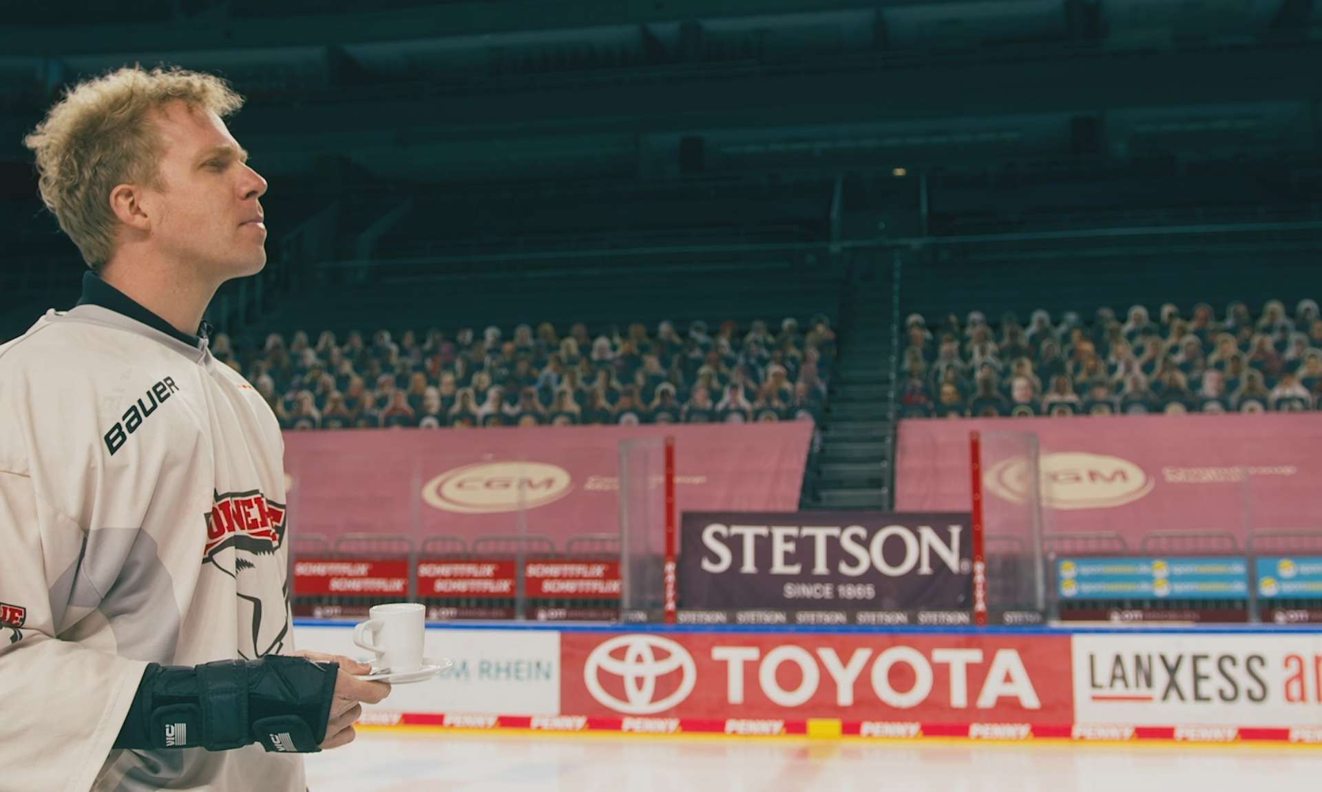 Ein Eishockeyspieler steht mit Kaffeebecher am Spielfeldrand einer Arena