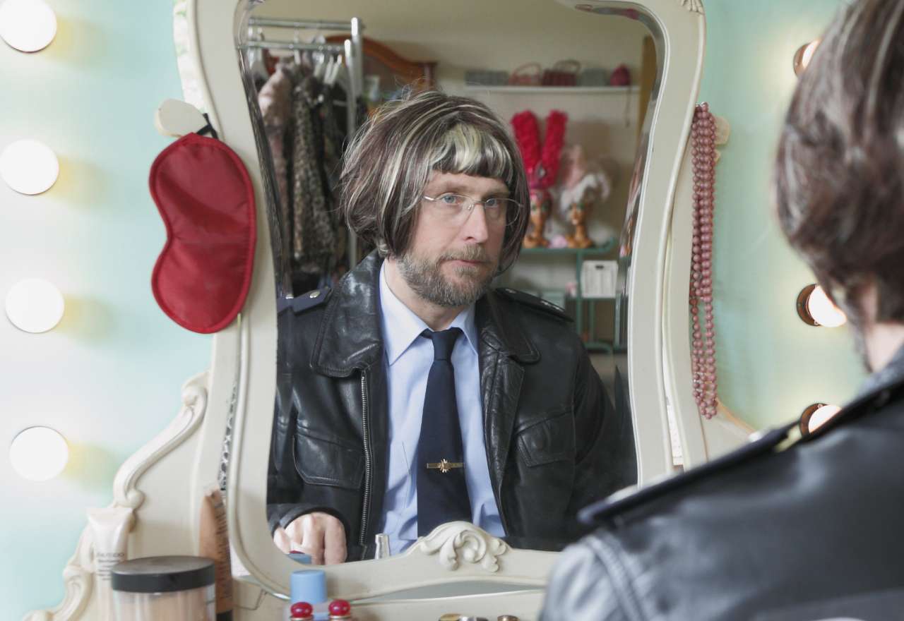 Ein Mann mit Perücke sitzt vor einem beleuchteten Garderobenspiegel
