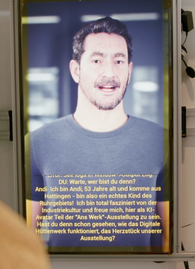 Bildschirmstele zeigt einen KI-Avatar, darunter läuft sein gesprochener Text mit
