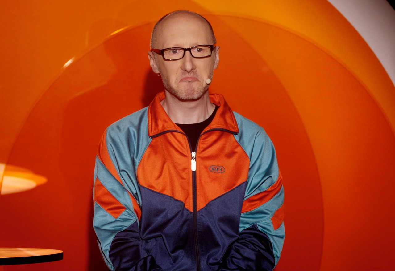 Ein Kabarettist steht vor einer orangefarbenen Bühnenwand